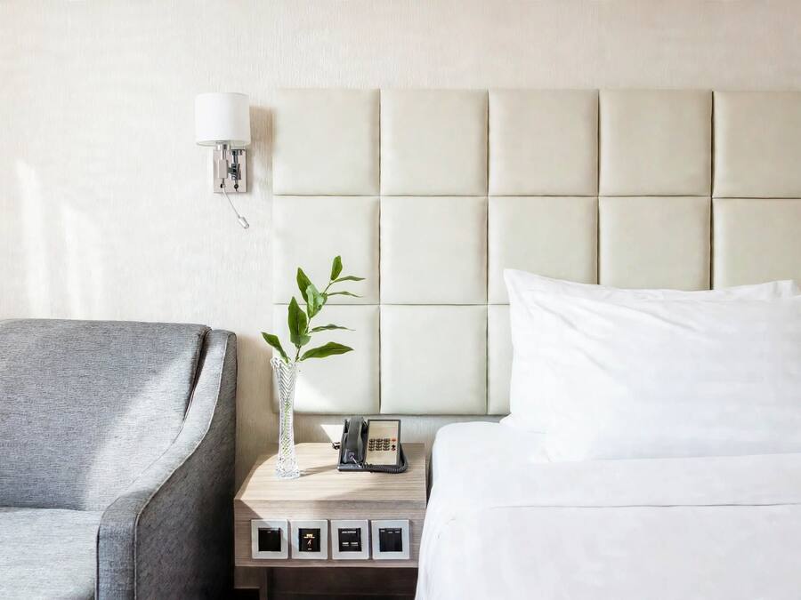Habitación de hotel con cama, mesita de noche con lámpara, teléfono y planta.