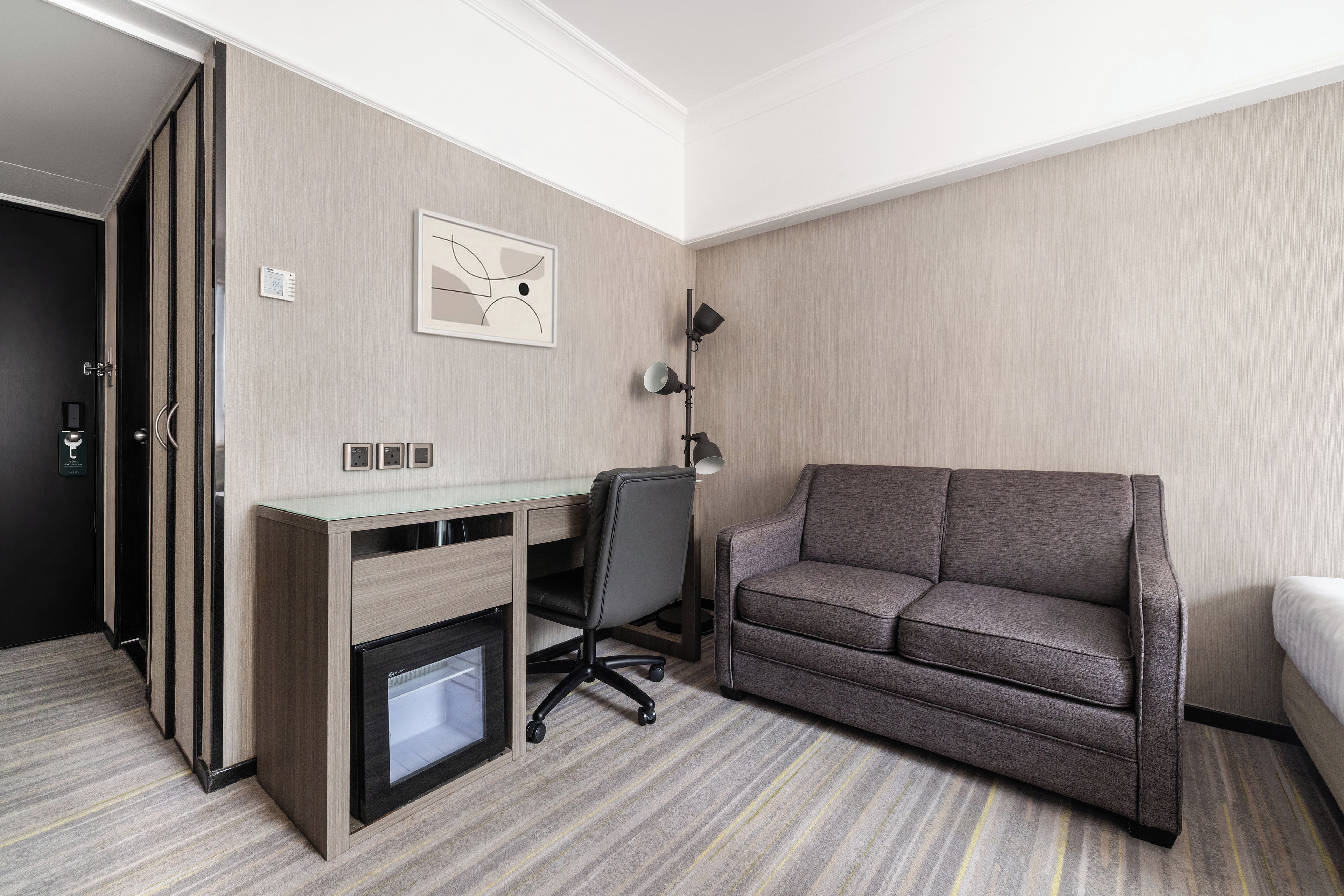 A hotel room with a desk, a chair, a sofa, a bed, and a wall-mounted lamp.