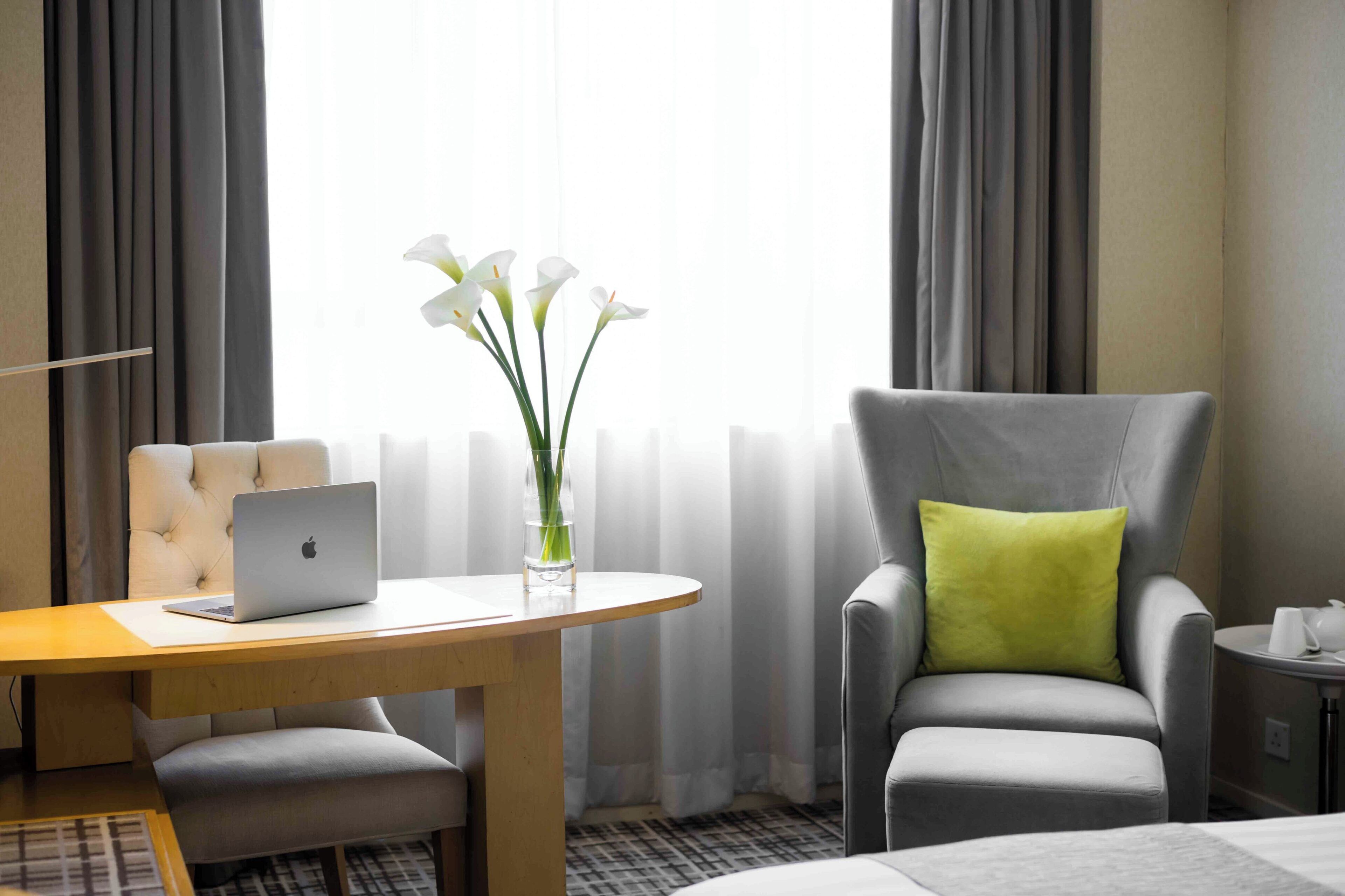 A hotel room with a bed, a desk with a laptop, a chair with a green pillow, and a vase with flowers.
