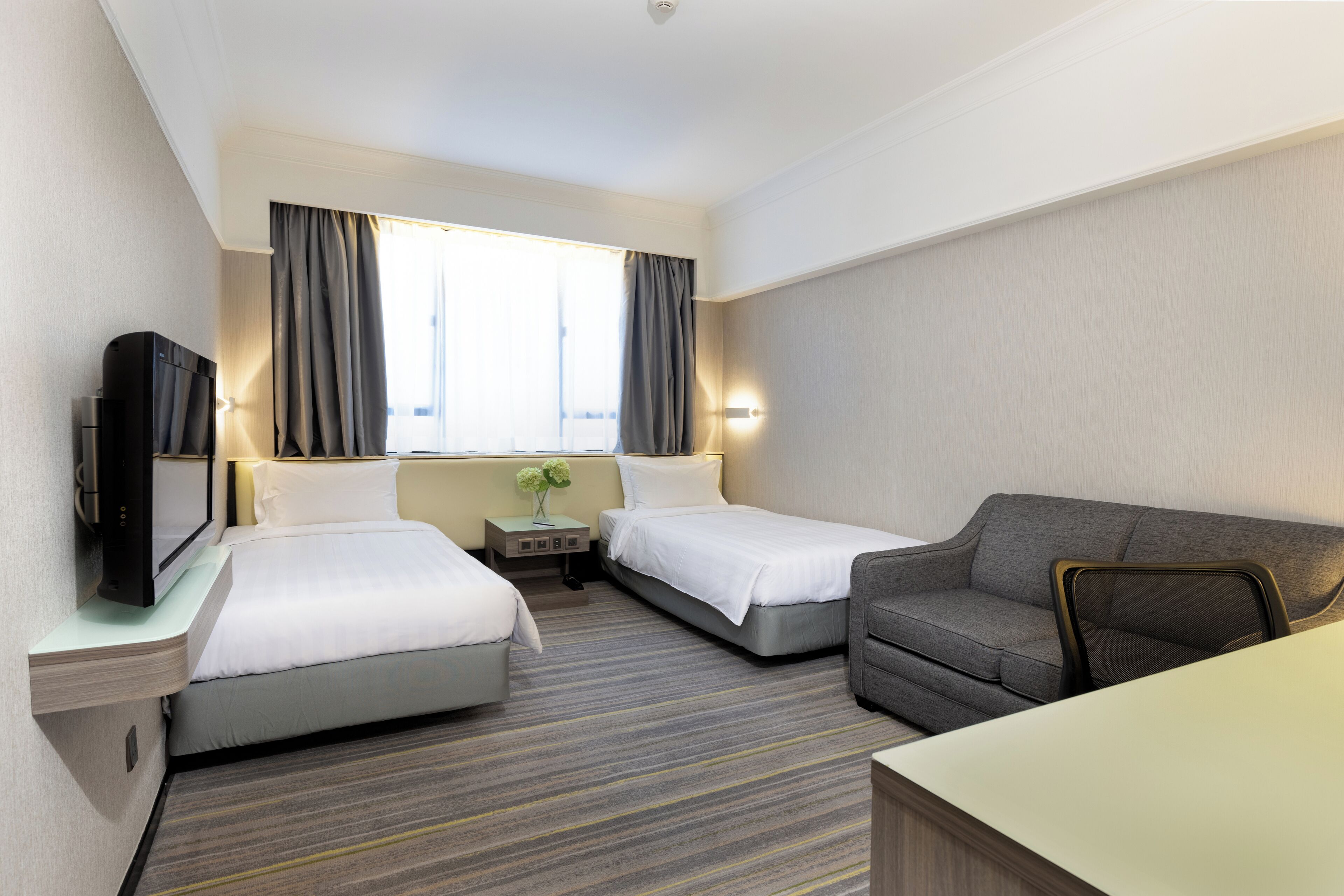A hotel room with two single beds, a sofa, a TV, a bedside table with a lamp, and a window with curtains.