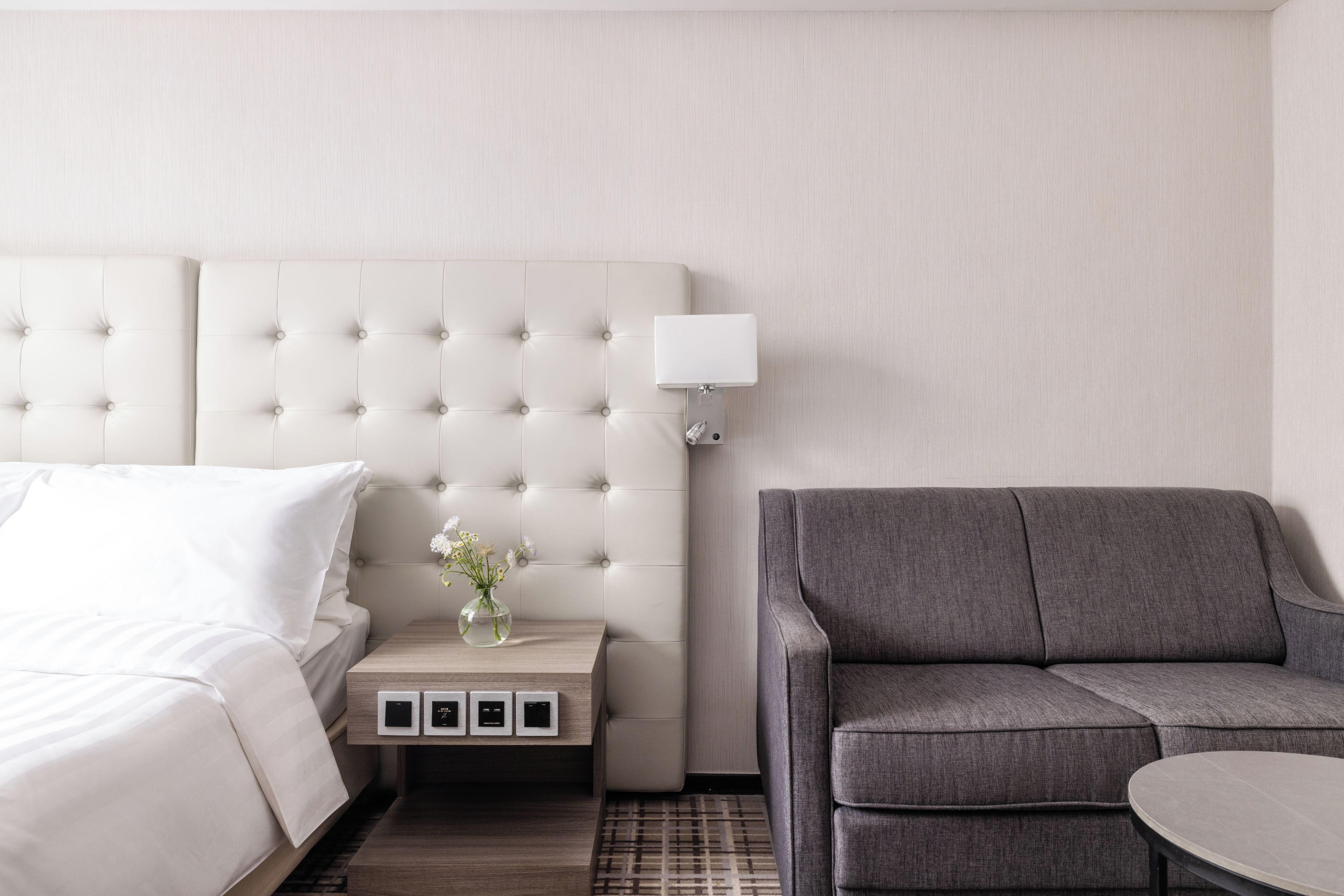 Et hotelværelse med en seng, et natbord med en lampe, en grå sofa og et lille bord med en blomstervase.