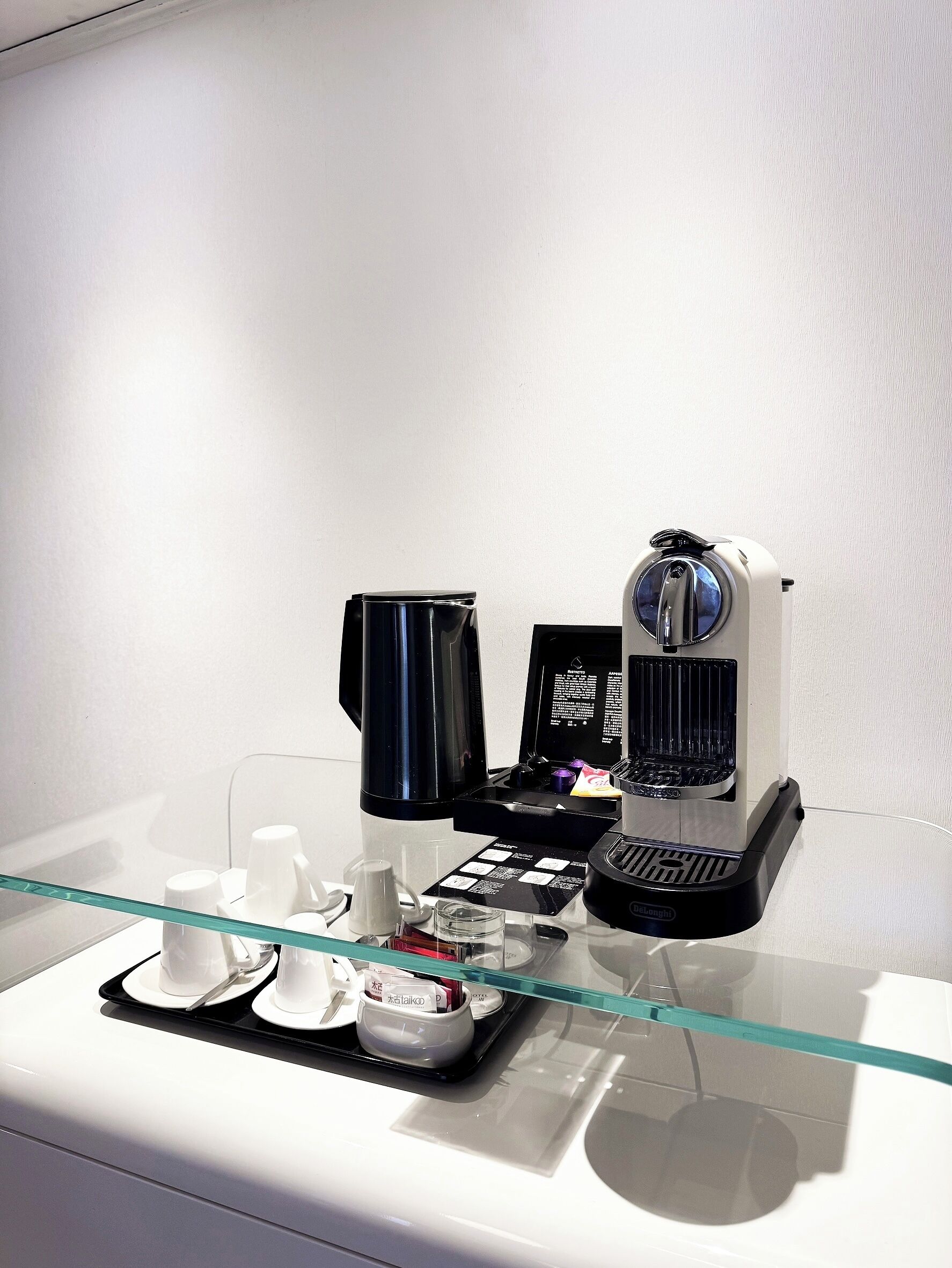 Máquina de café sobre uma mesa de vidro, com uma bandeja de copos brancos e um recipiente preto.