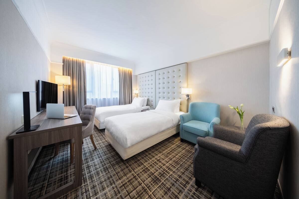 A hotel room with two beds, a desk with a laptop, a chair, and a small table with a vase of flowers.