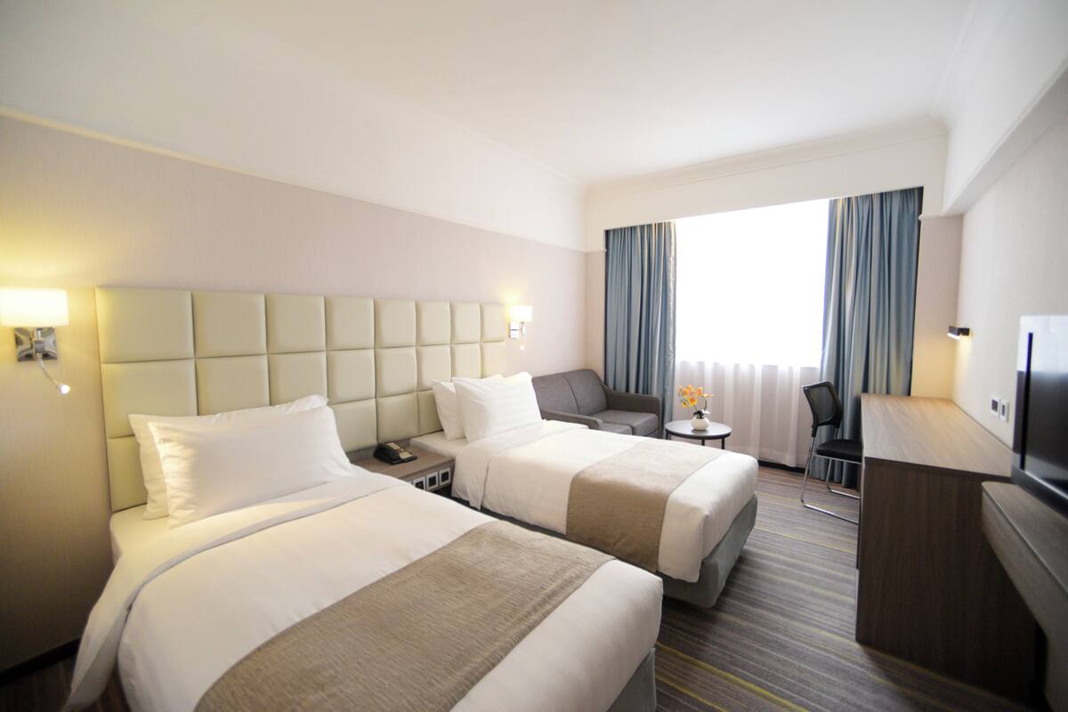 A hotel room with two beds, a desk, a chair, a TV, and a window with curtains.