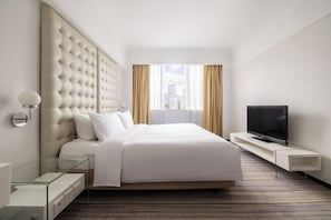 Una habitación de hotel con una cama grande, un televisor de pantalla plana, una mesita de noche de vidrio y vistas a la ciudad desde la ventana.
