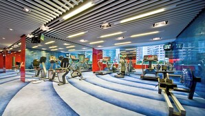 A modern gym with various cardio machines, a blue carpeted floor, and a cityscape view through large windows. - Panda Hotel (Tsuen Wan)
