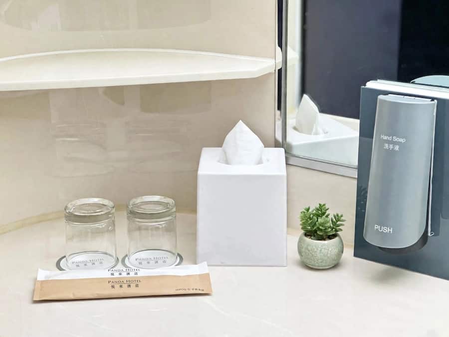 Un baño con dispensador de jabón, caja de pañuelos, dos botellas de vidrio, una pequeña planta en maceta y dispensador de toallas.