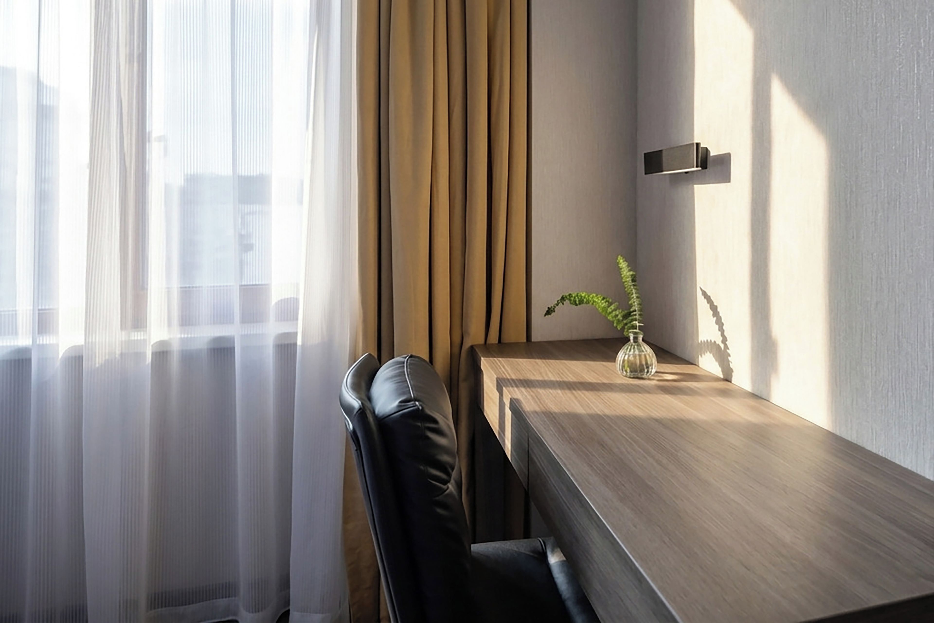 A modern hotel room with a wooden desk, a chair, a potted plant, and sheer curtains.