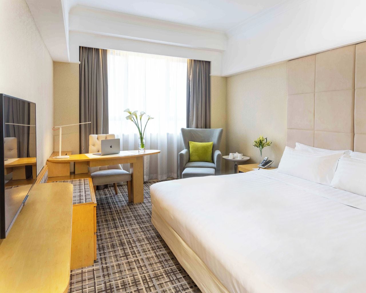 A hotel room with a large bed, a desk with a laptop, a chair, a small table with a phone, and a TV.