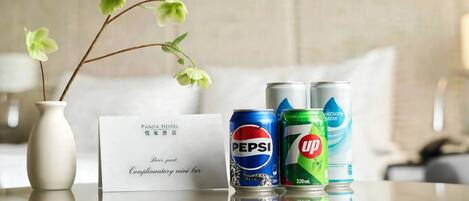 Habitación de hotel con un jarrón blanco con flores, un minibar de cortesía y dos latas de bebida.