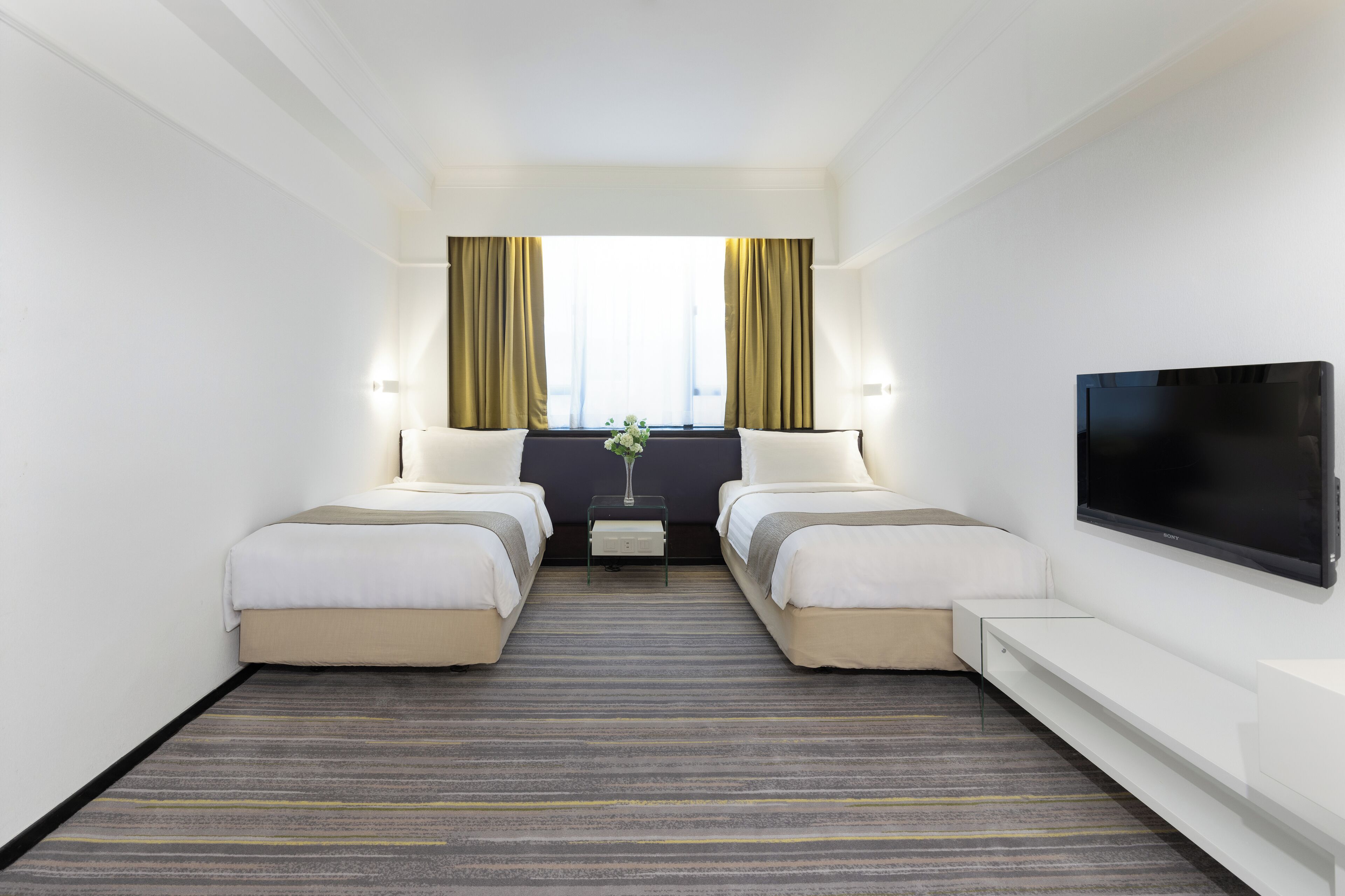 Quarto de hotel com duas camas, televisão de tela plana, janela com cortinas e uma mesinha com um vaso.