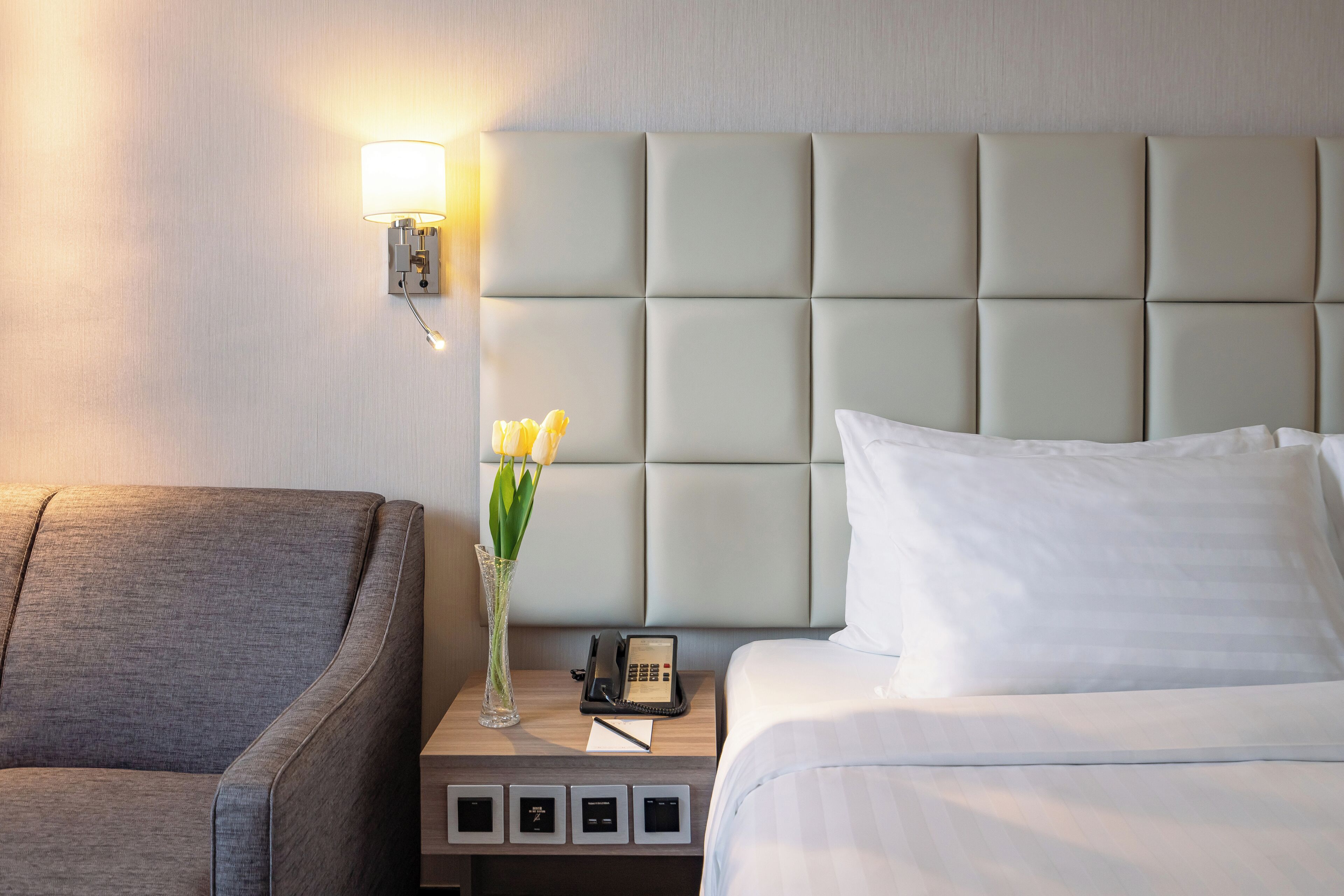Ett hotellrum med en stoppad sänggavel, ett nattduksbord med en lampa, en vas med blommor, en telefon och en säng med vita sängkläder.