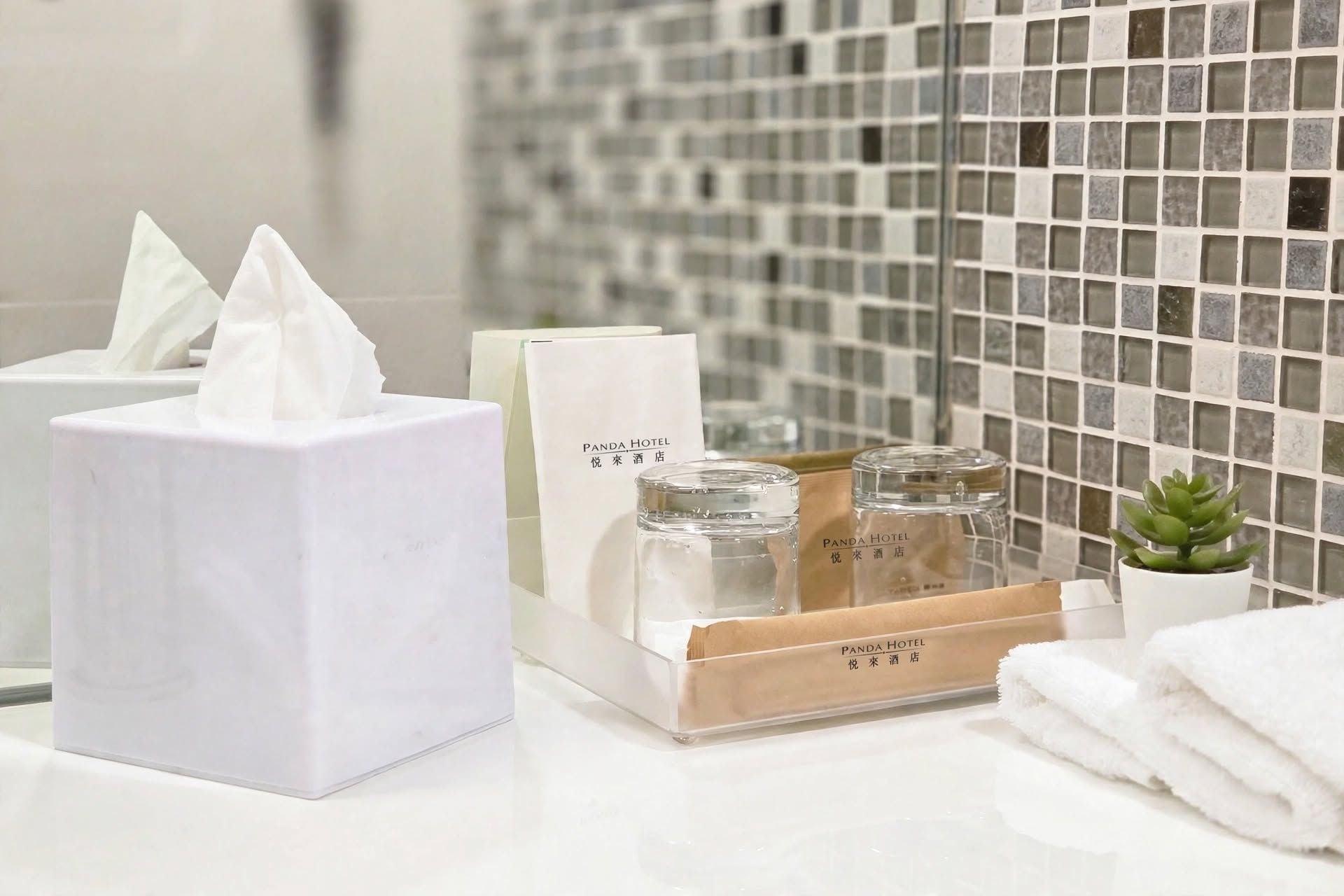 Una encimera de baño con una caja de pañuelos, dispensador de pañuelos, una alfombrilla de baño de bambú, una pequeña planta en maceta y un frasco de vidrio con etiqueta.