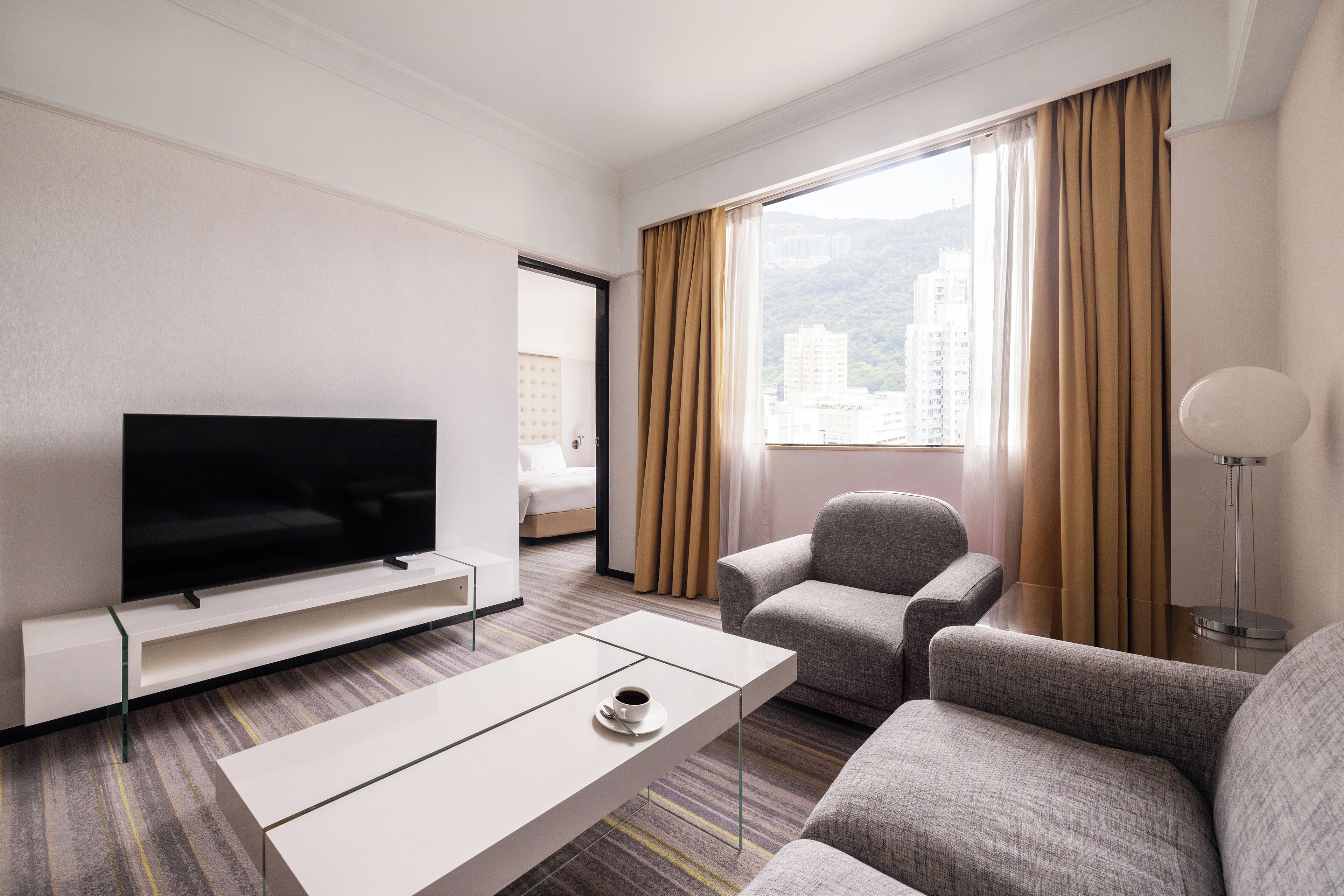 A hotel room with a flat-screen TV, a sofa, a chair, a coffee table, and a view of buildings through the window.