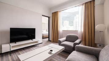 A hotel room with a flat-screen TV, a sofa, a chair, a coffee table, and a view of buildings through the window.