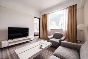 Quarto de hotel com televisão de tela plana, sofá, poltrona, mesa de centro e vista para edifícios pela janela.