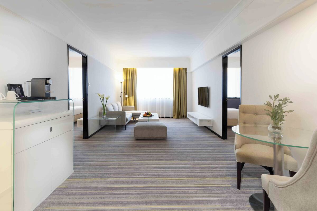 A modern hotel room with a glass desk, a sofa, a coffee machine, and a TV on the wall.