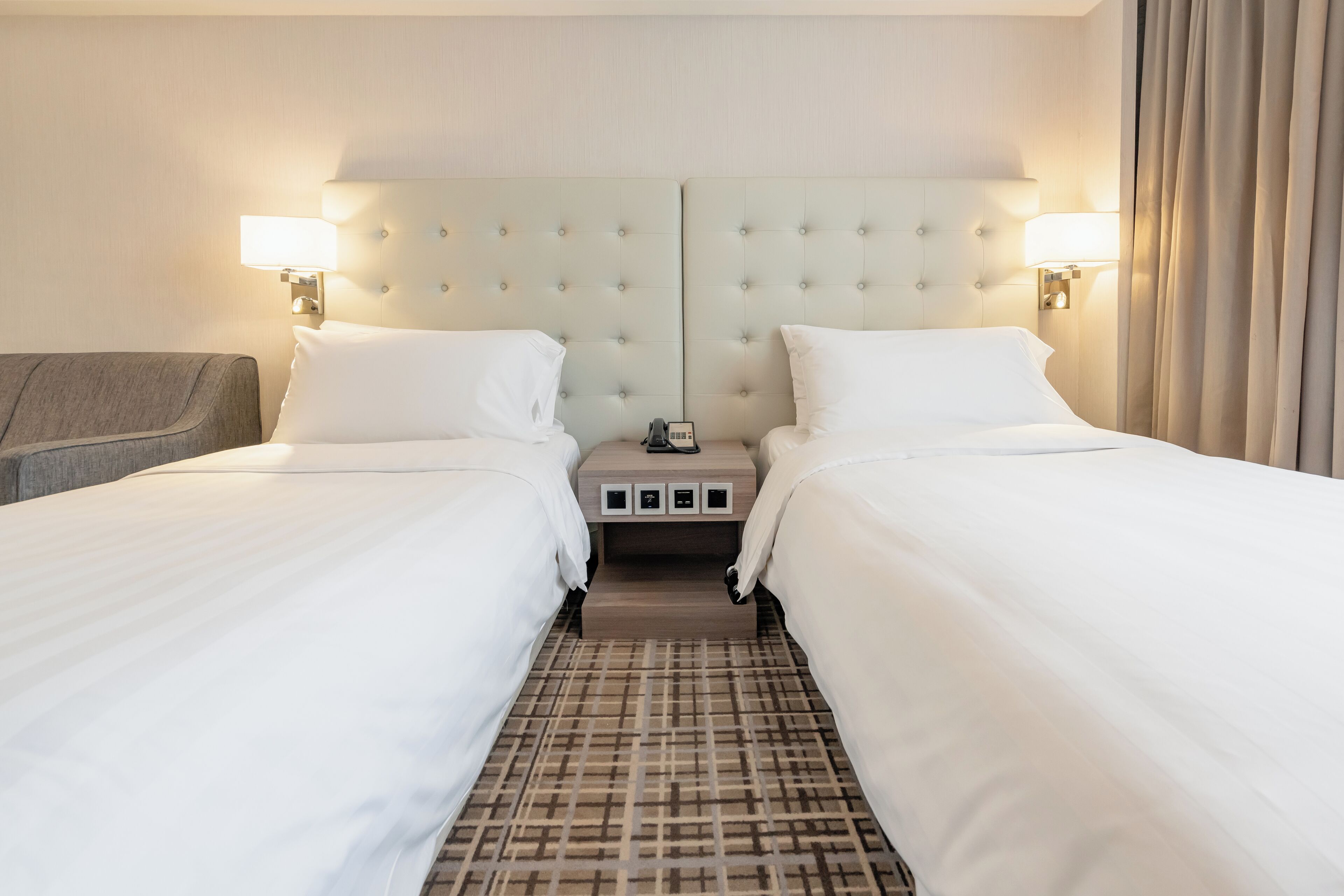 A hotel room with two beds, a headboard with a mirror, bedside lamps, a nightstand, and a patterned carpet.