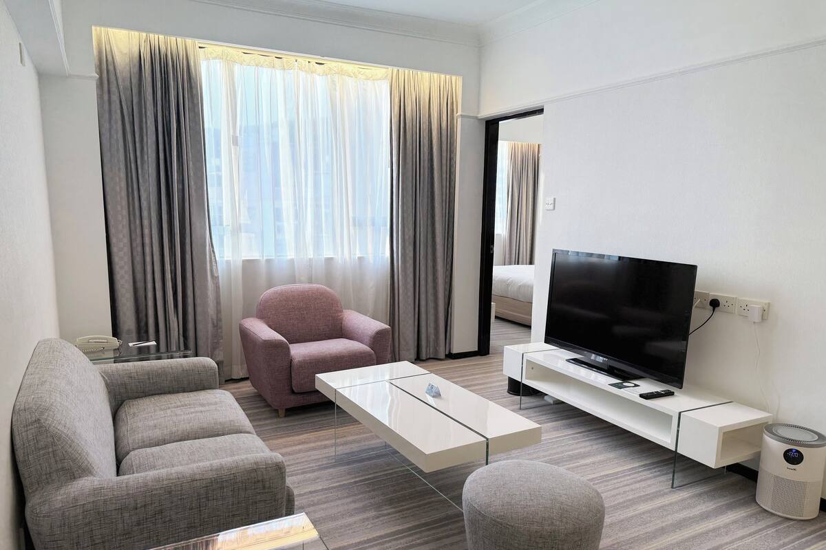 A modern hotel room with a sofa, armchair, TV, and a small ottoman.