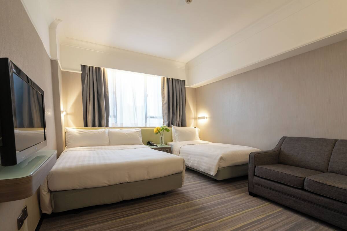 A hotel room with two beds, a TV, a sofa, and a window with curtains.
