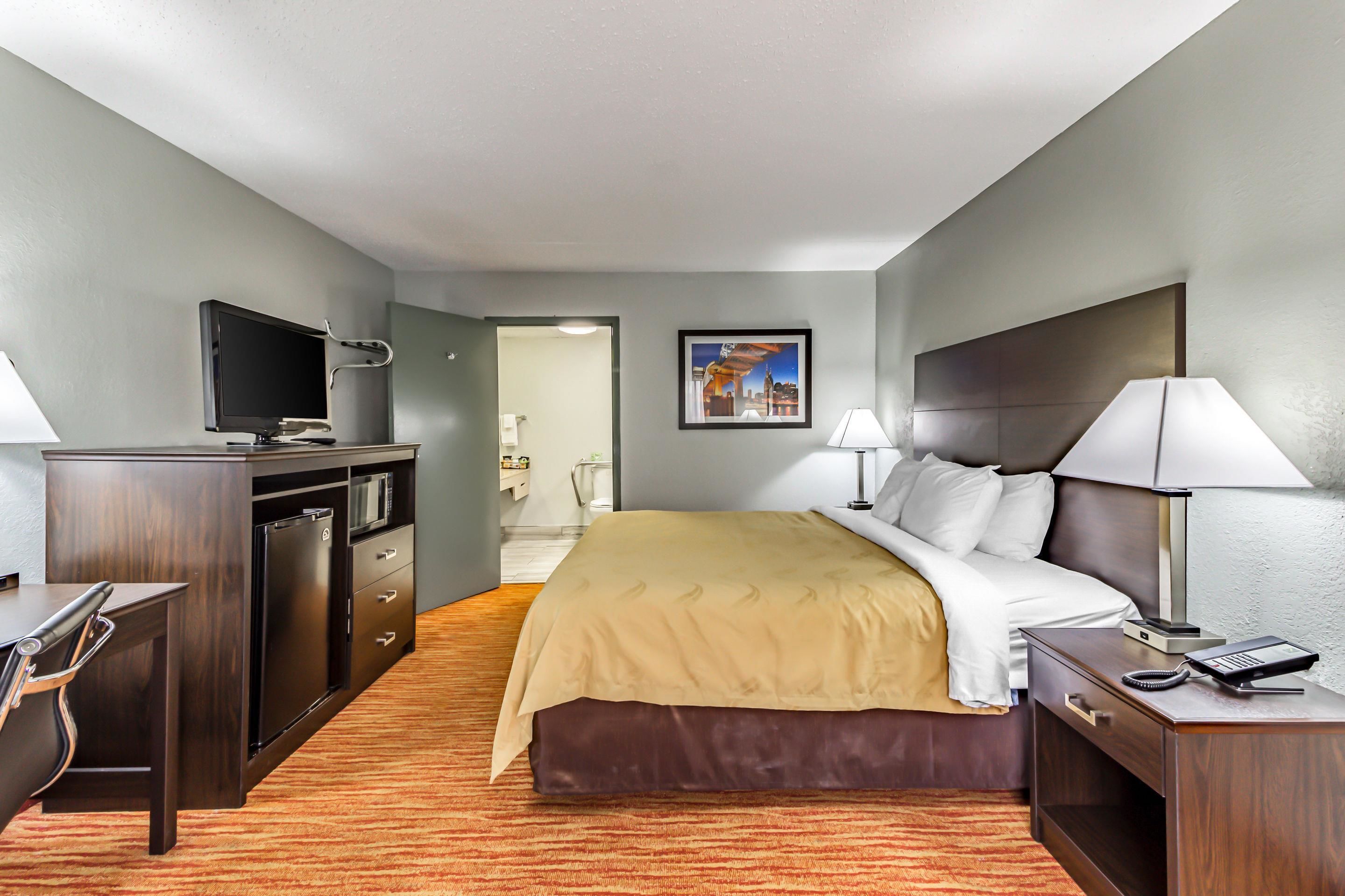 A hotel room with a bed, bedside table, lamp, desk, TV, and a view of the hallway.