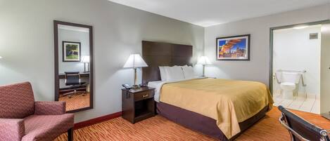 Habitación de hotel con cama, dos lámparas de noche, una silla, un escritorio con silla, un espejo y un cuadro en la pared.