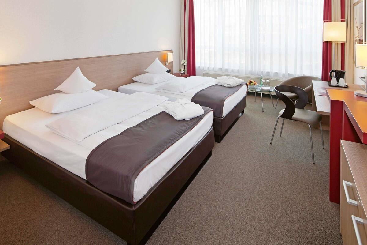 A hotel room with two beds, a desk, a chair, and a window with curtains.