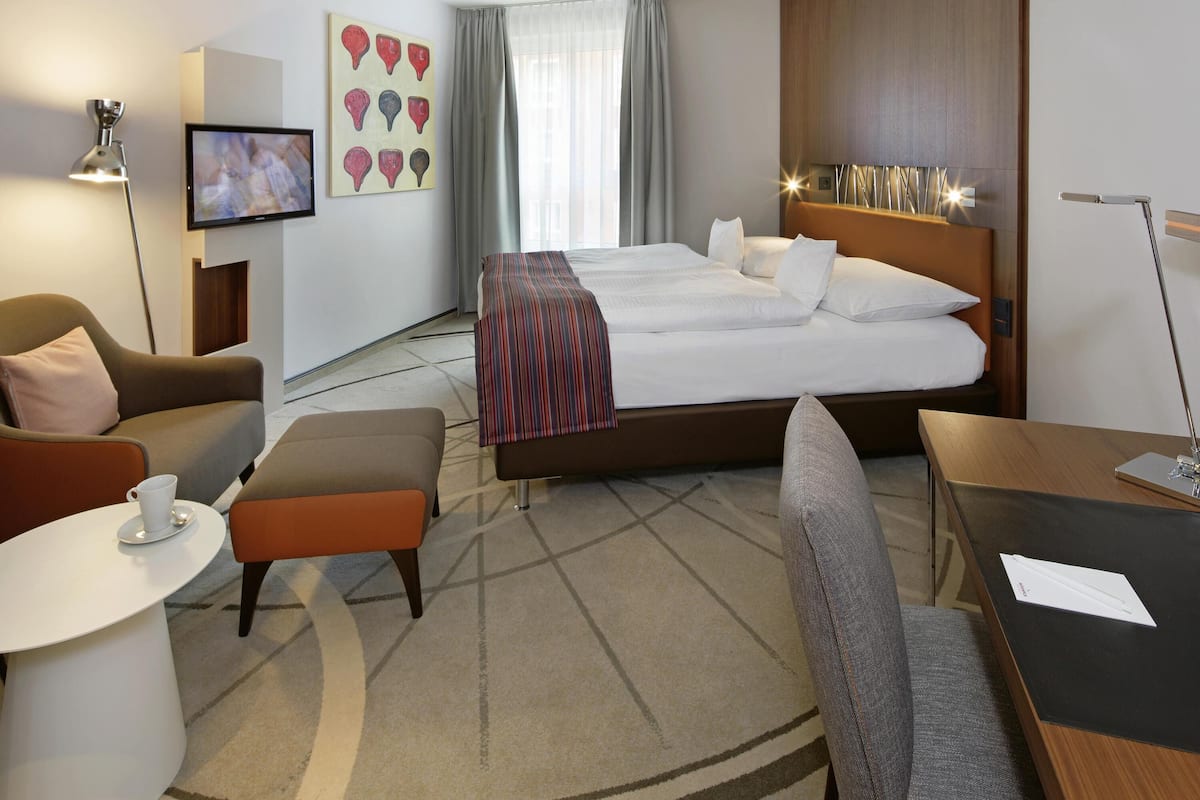A hotel room with a bed, a desk, a chair, a sofa, a TV, and a painting on the wall.