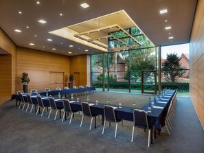 A conference room with a long table, chairs, and a large window with a view of greenery and buildings. - Mövenpick Hotel Münster am Aasee (Münster)