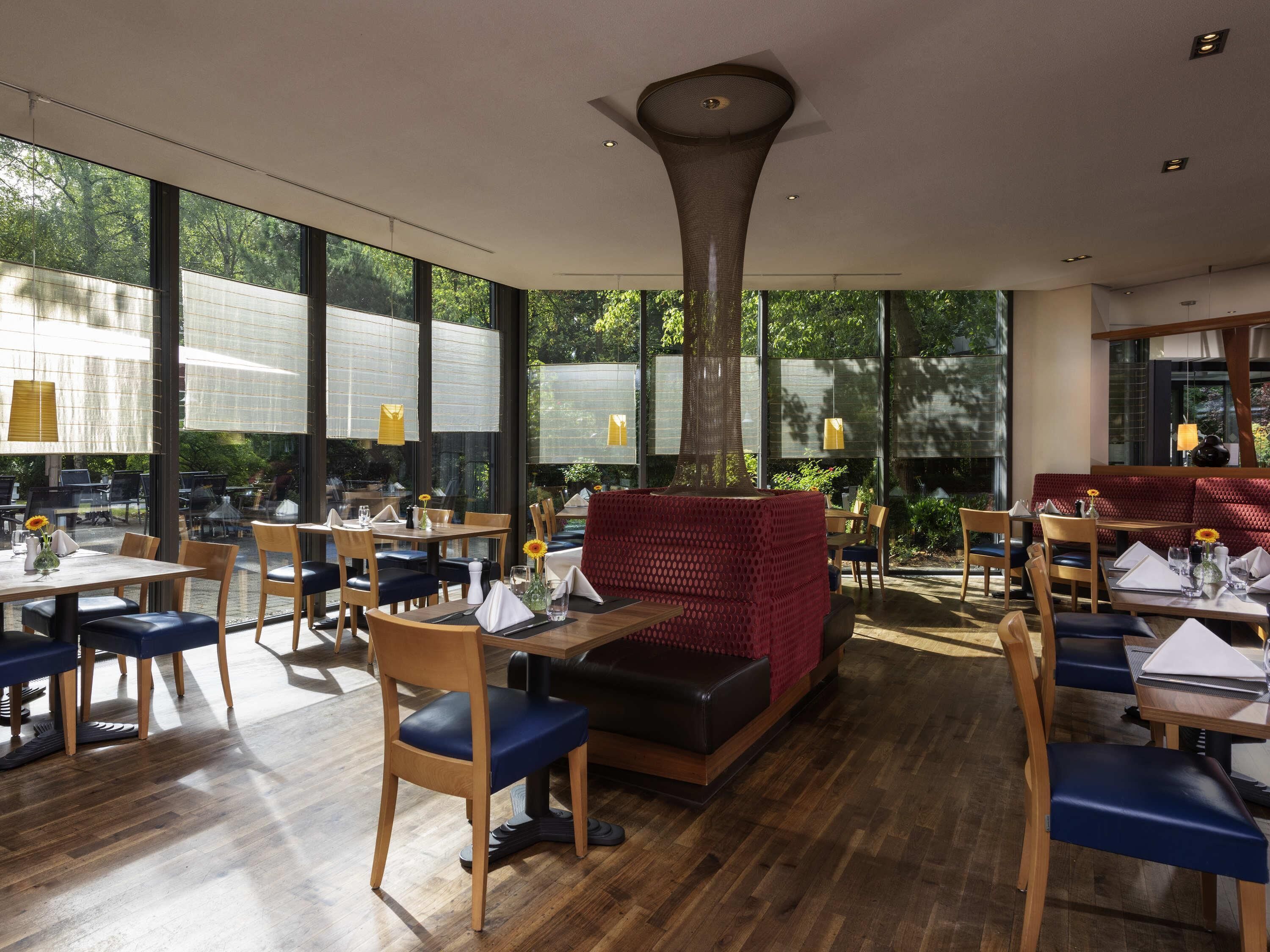 Un restaurante con amplios ventanales, mesas y sillas de madera, y una zona de estar central roja.