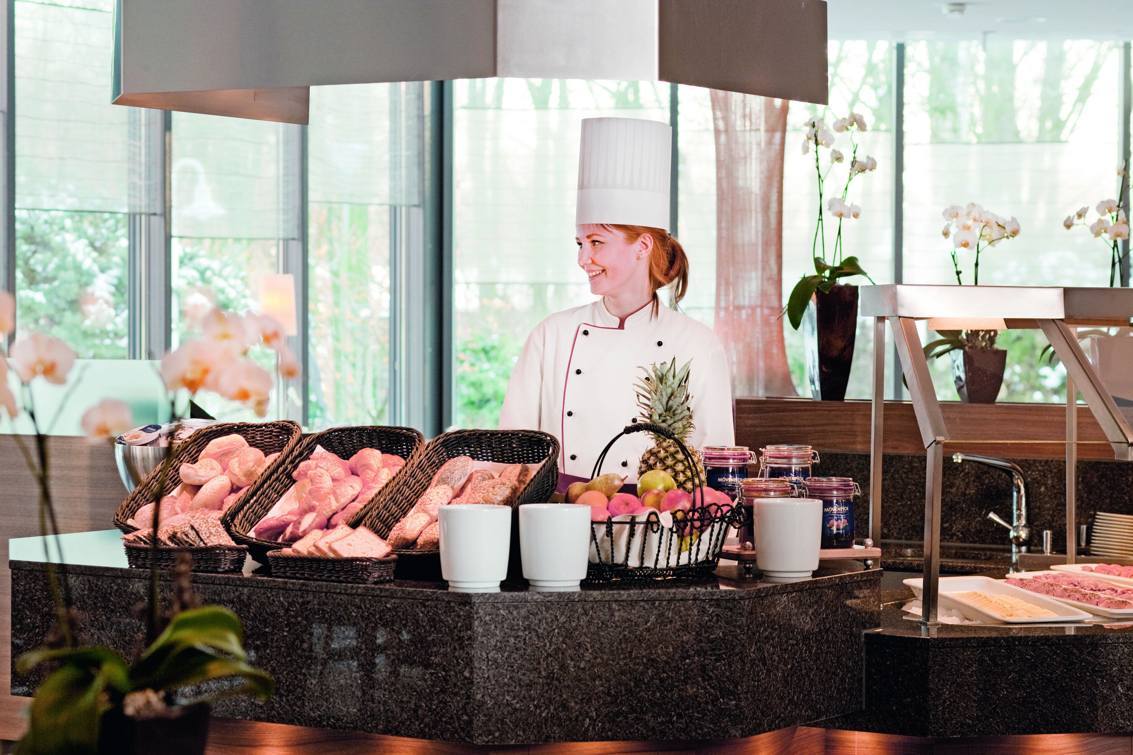 Um chef com uniforme branco e um chapéu alto, de pé atrás de um balcão de buffet com cestas de pão e frutas.