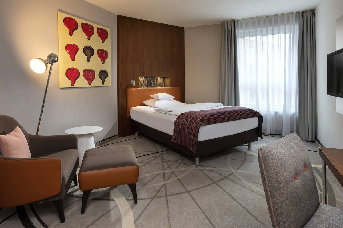 A hotel room with a bed, a desk, a chair, a lamp, and a painting on the wall.