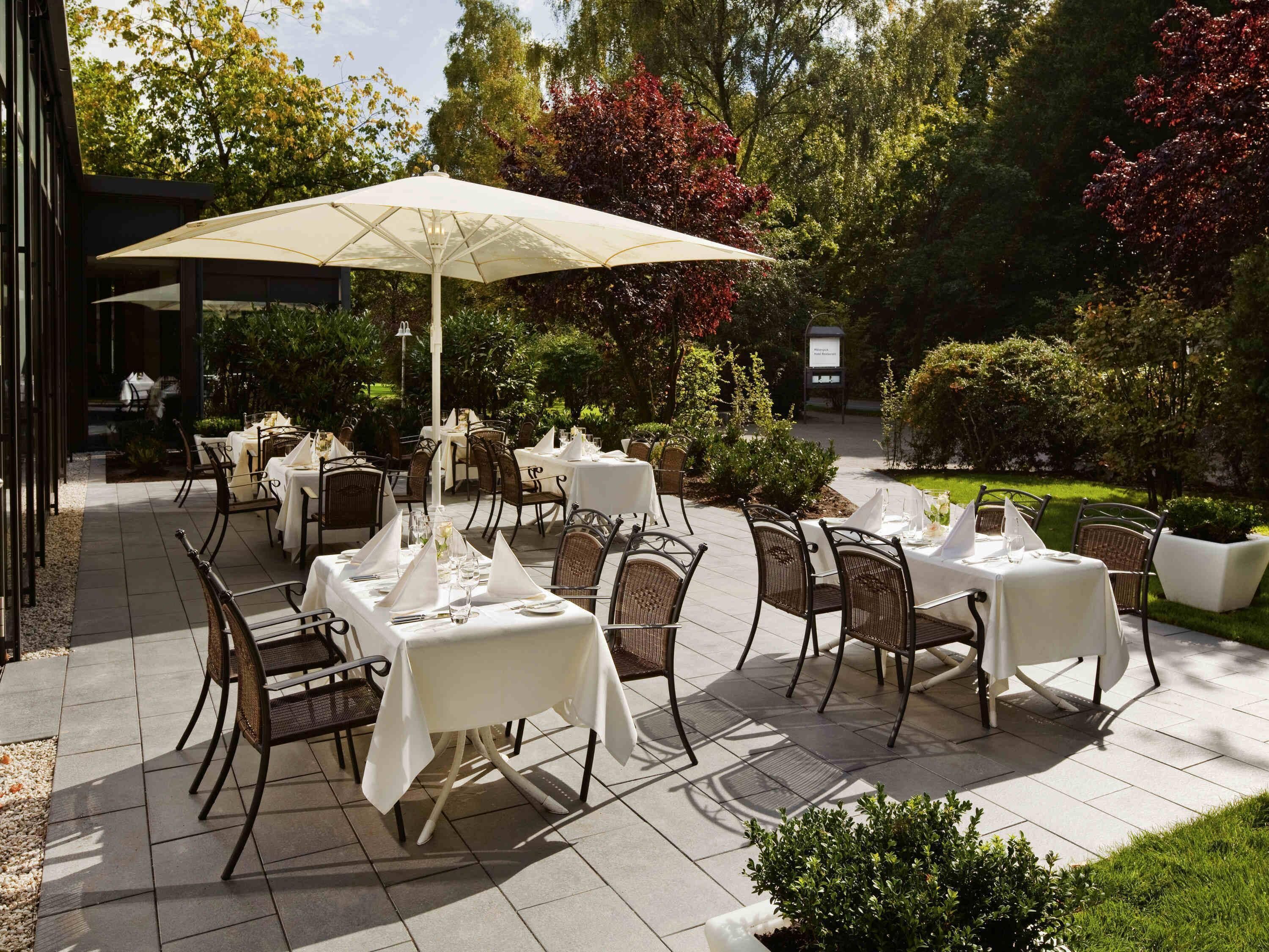 An outdoor patio with tables and chairs, surrounded by greenery and trees.