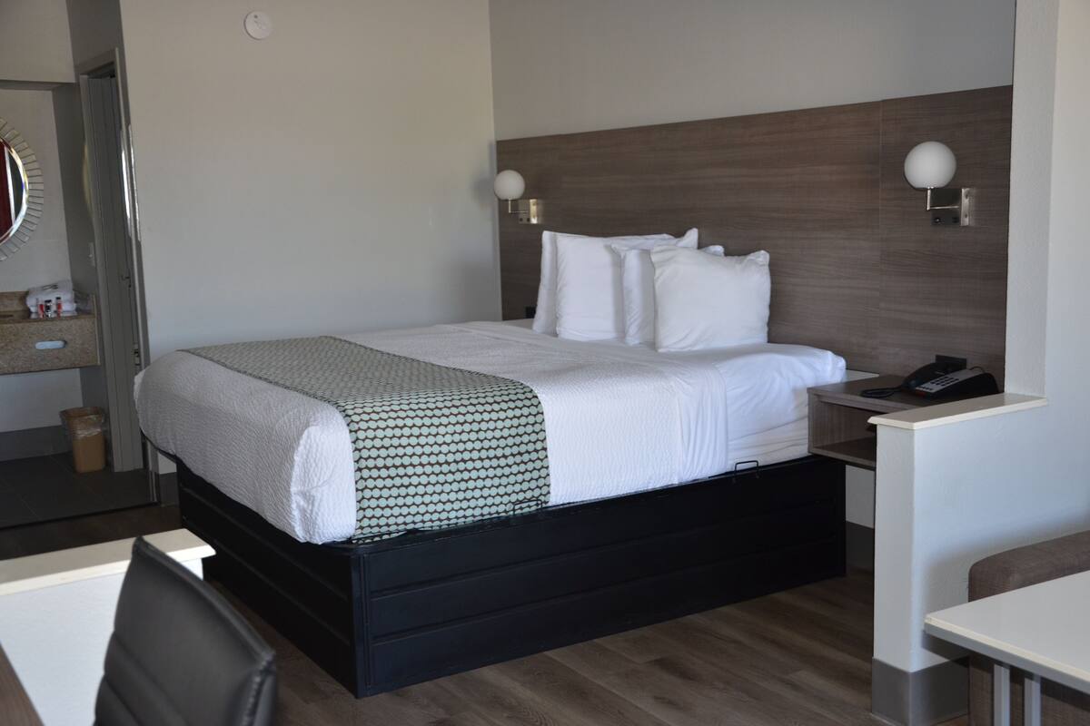Cama bem arrumada com lençóis brancos e manta estampada, mesa de cabeceira com telefone e luminária fixada na parede.