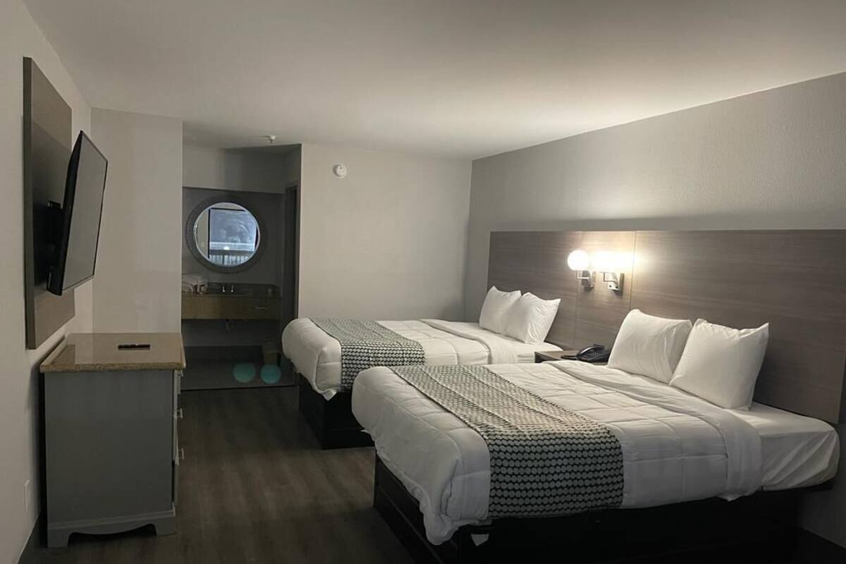 Quarto de hotel com duas camas, televisão, secretária pequena, janela redonda e cabeceira com iluminação.