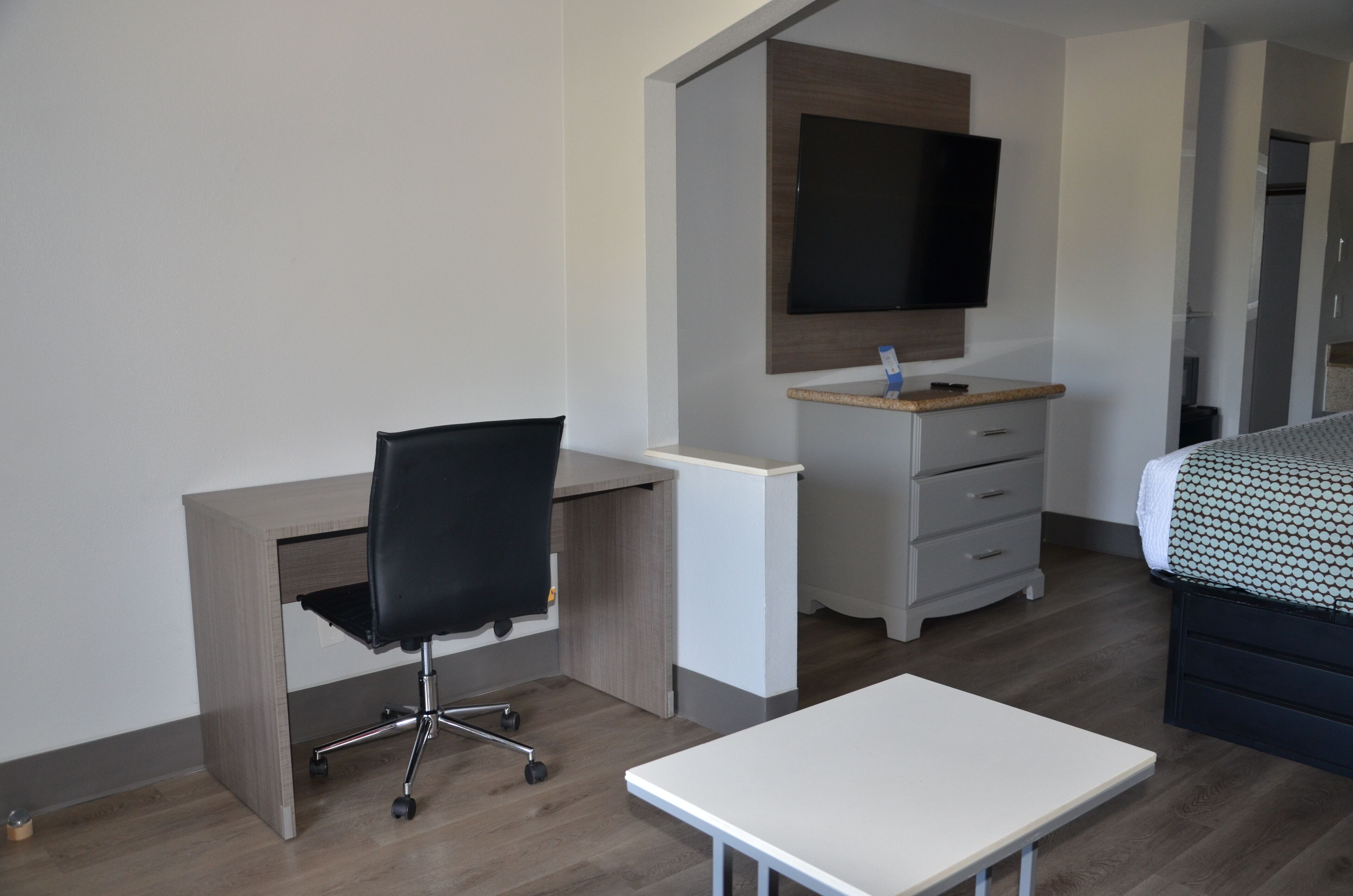A modern hotel room with a wooden desk, black office chair, a flat-screen TV mounted on a wooden panel, a white bedside table, and a bed with a patterned bedspread.