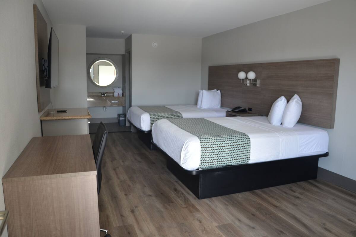 Quarto de hotel com duas camas, uma escrivaninha, um espelho e piso de madeira.