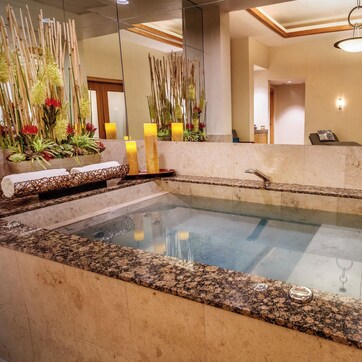 A spa area with a glass-enclosed hot tub, a wooden bench with towels, and a decorative plant arrangement.