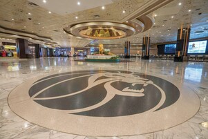 En rymlig entréhall med ett stort, cirkelformat emblem på golvet, en centralt placerad dekorativ lampa och en digital skärm som visar innehåll.