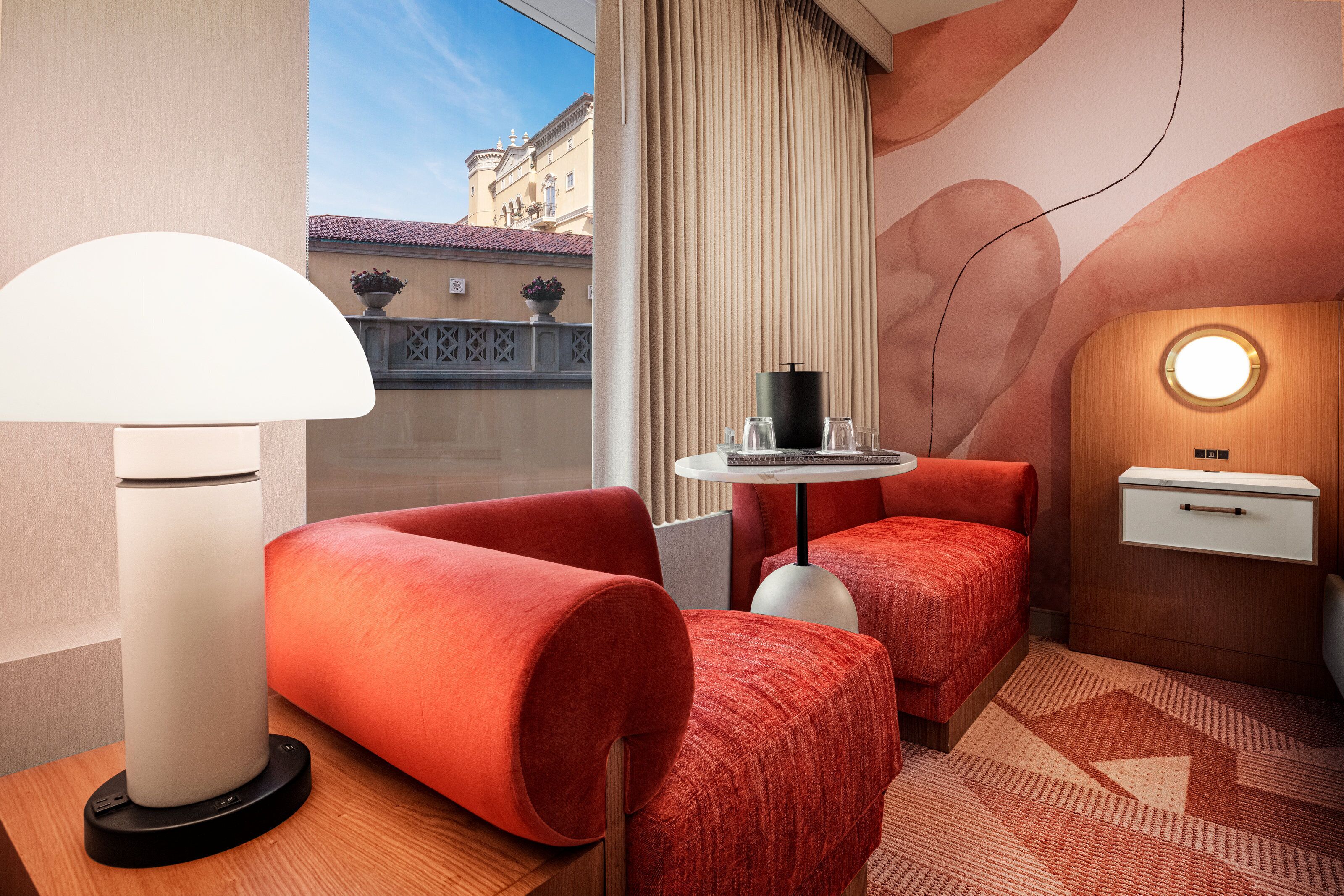 A hotel room with a bed, a desk, a lamp, and a view of the outside.