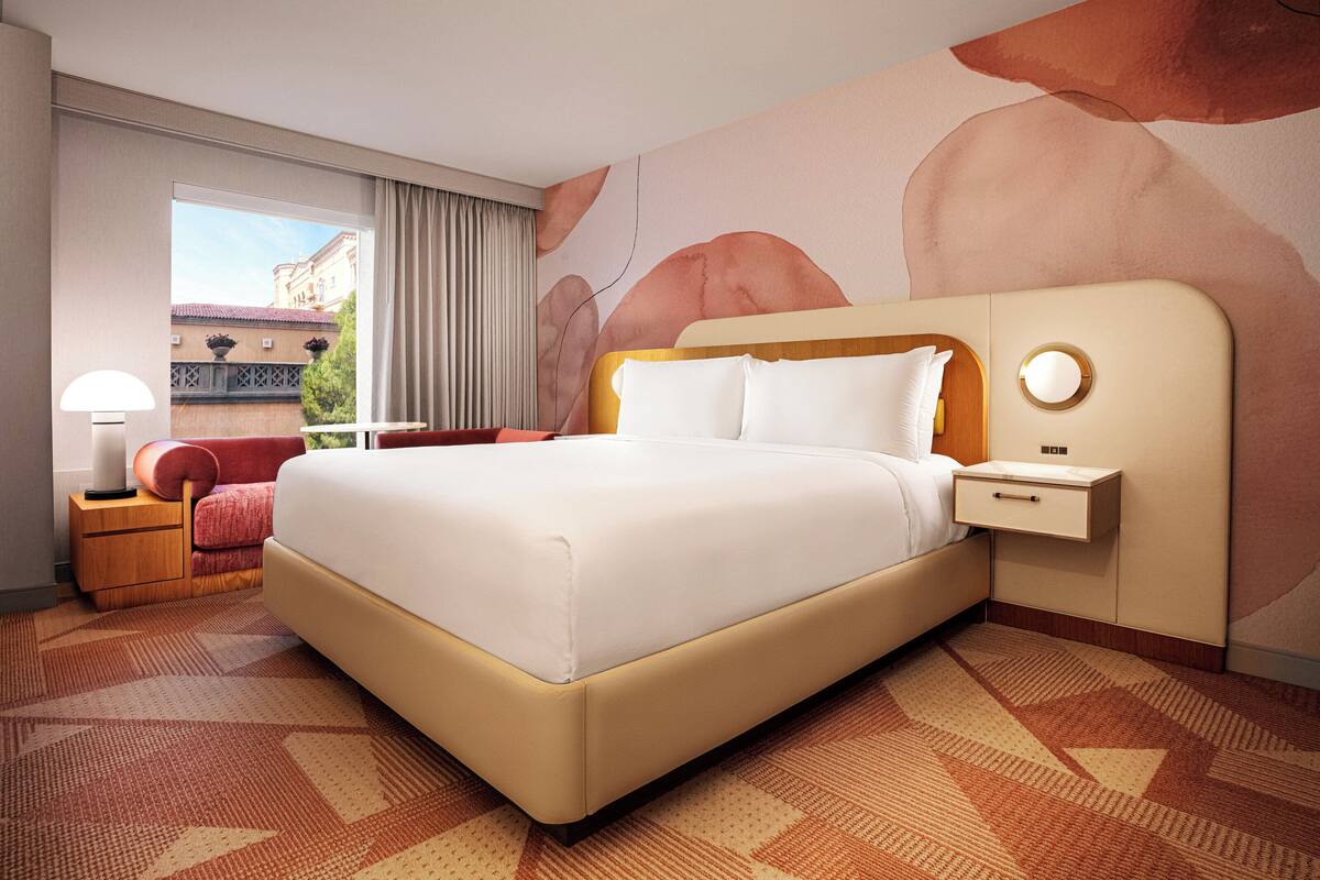 A hotel room with a large bed, a bedside table with a lamp, a nightstand, and a view of a building outside.