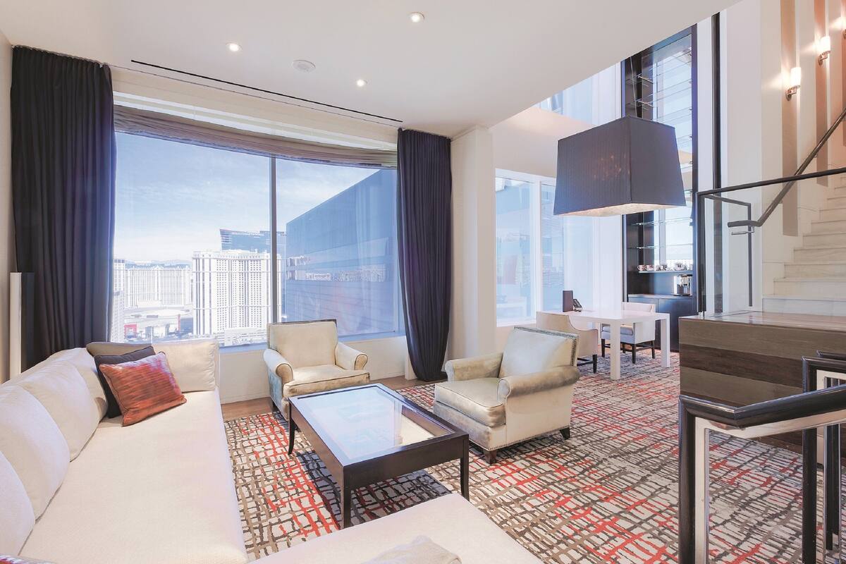 A modern hotel room with a large window, a sofa, a coffee table, and a staircase.