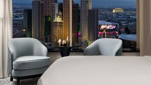 A modern hotel room with a large window offering a cityscape view, two armchairs, a small table, and a decorative vase.