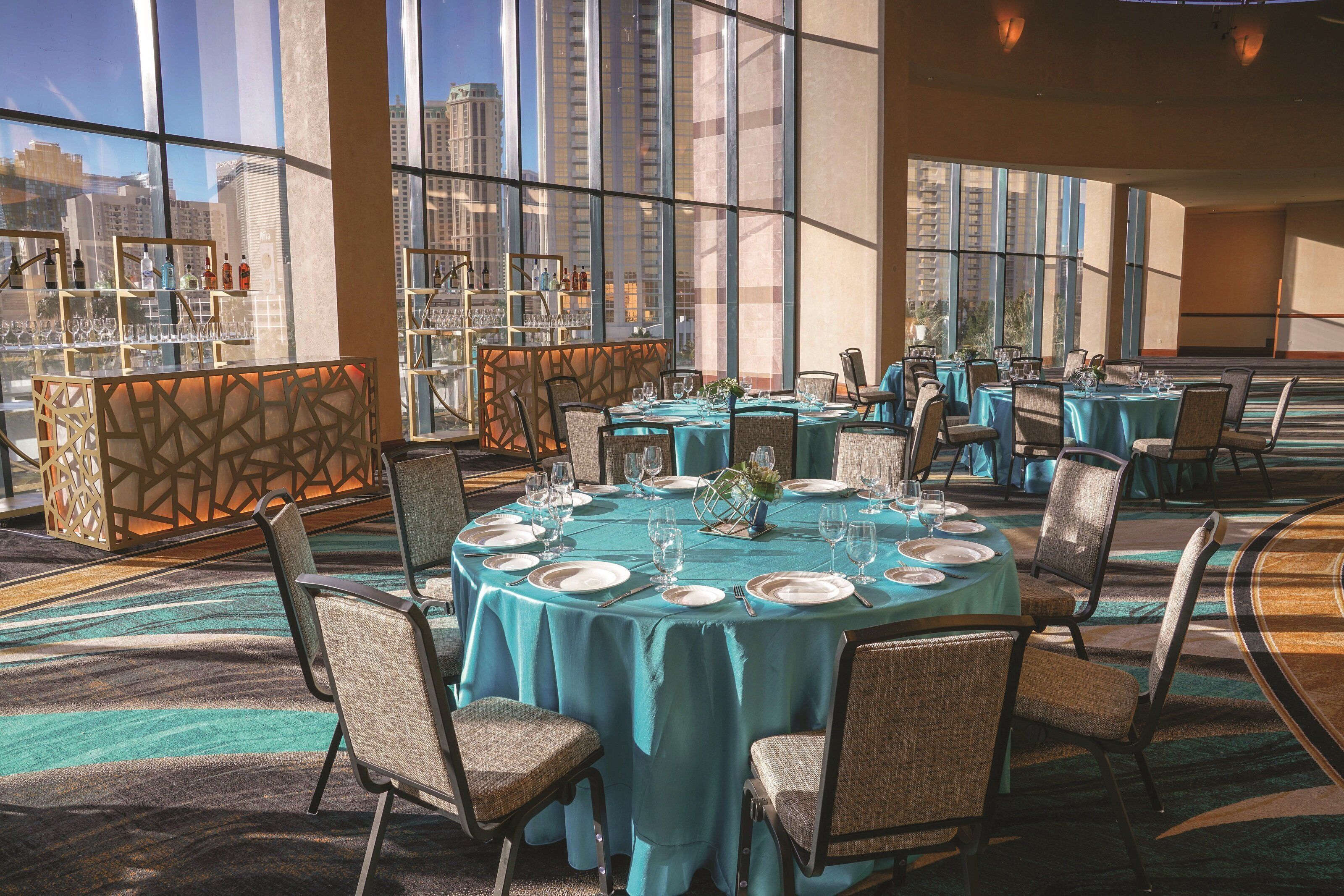 Un restaurant avec des tables rondes disposées pour le service, de grandes fenêtres offrant une vue sur la ville et un bar avec des étagères remplies de bouteilles.