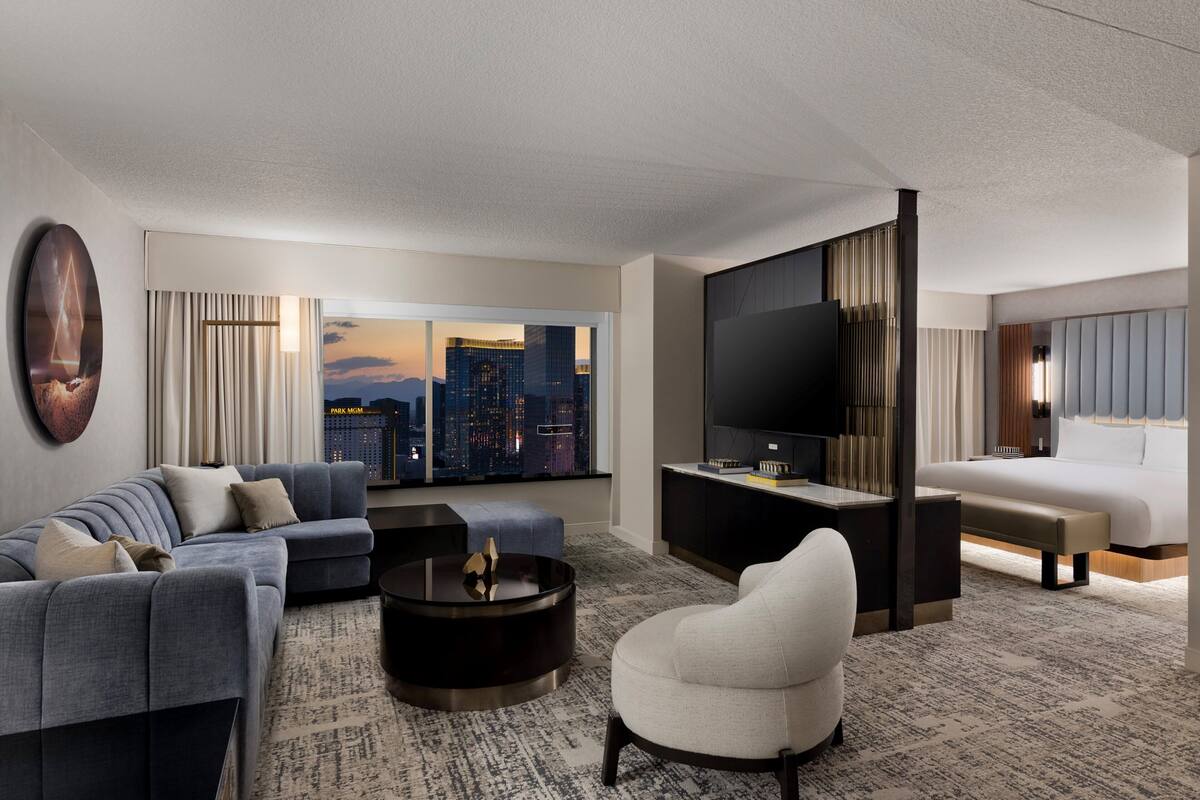 A modern hotel room with a city view, a large flat-screen TV, a sofa, a chair, and a bed.