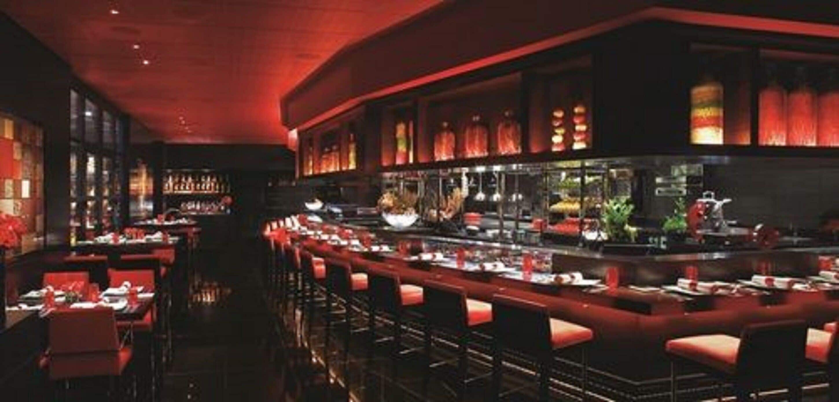 A dimly lit restaurant with red ambient lighting, multiple tables, and a bar area.