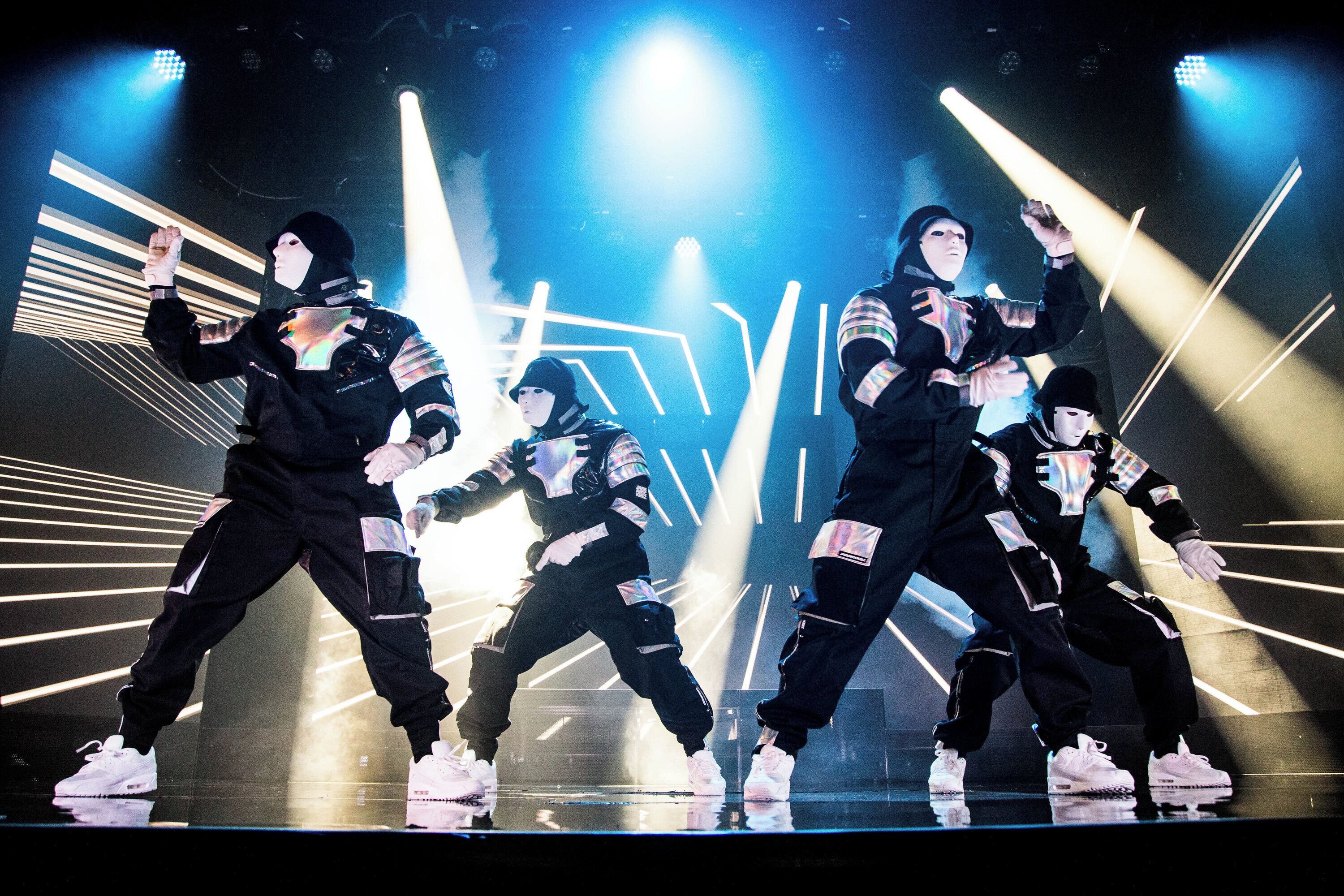 Un grupo de bailarines actuando en un escenario con luces brillantes y patrones geométricos de fondo.