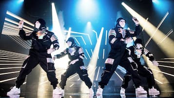 Un grupo de bailarines actuando en un escenario con luces brillantes y patrones geométricos de fondo.