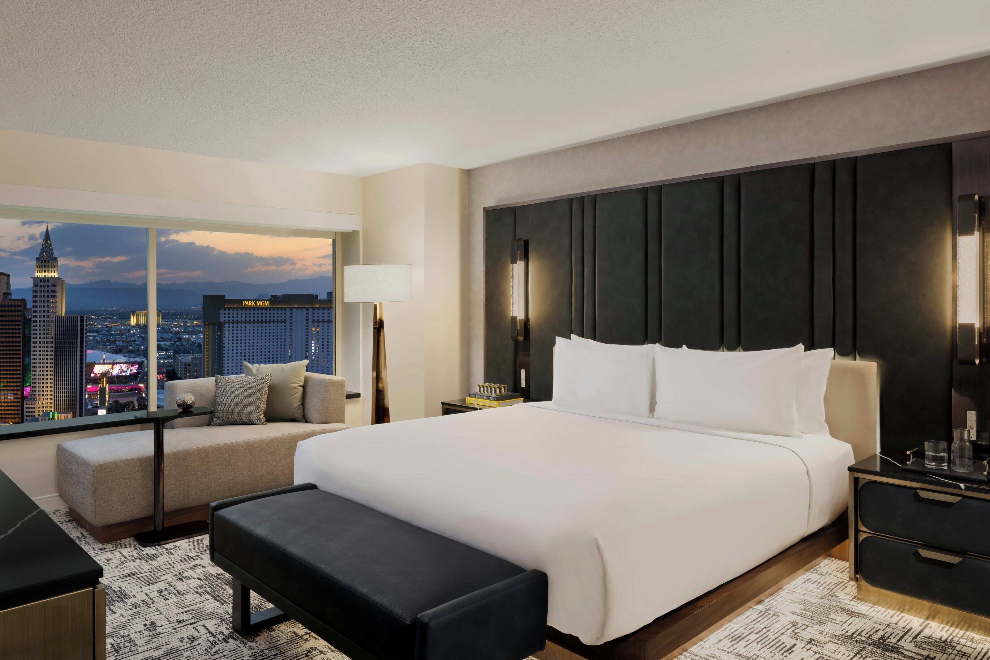 A modern hotel room with a large bed, a sofa, and a desk. There is a view of the cityscape through large windows.