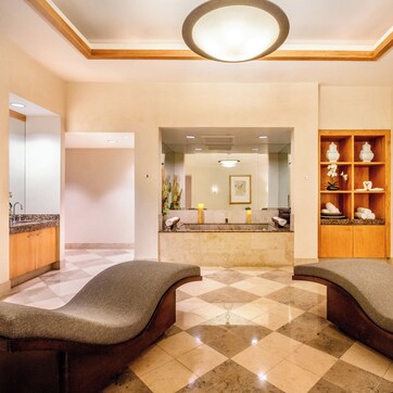 A hotel lobby with a checkered floor, two cushioned seating areas, a reception desk, and a wooden shelving unit with decorative items.